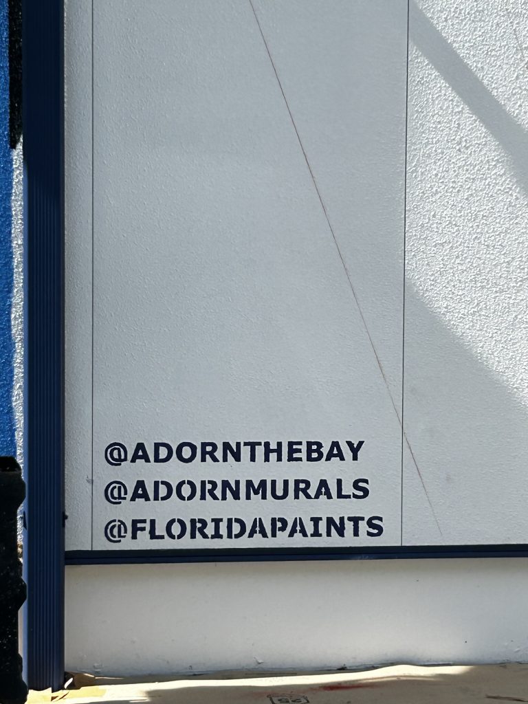 A close-up of a white wall with the social media handles of Adorn the Bay and Florida Paints.
